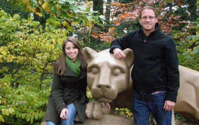 An adult brother and sister pose with the Penn State University Nittany Lion statue. It is autumn and they are wearing coats.