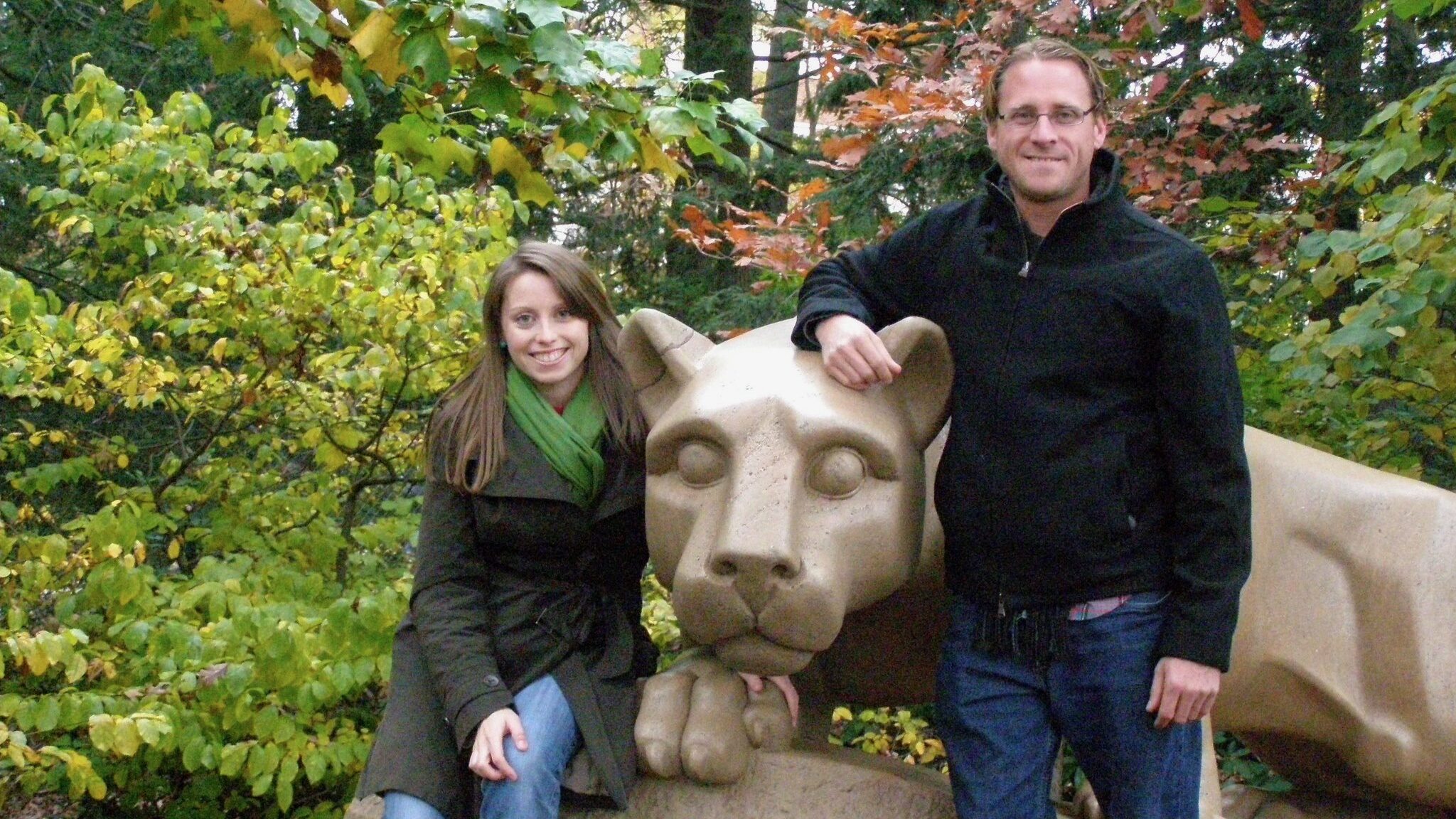 An adult brother and sister pose with the Penn State University Nittany Lion statue. It is autumn and they are wearing coats.