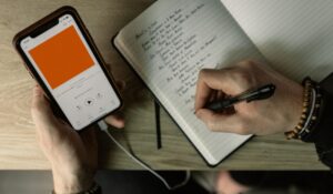 A person holds an iPhone that shows a podcast playing. They are taking notes in a notebook while listening.