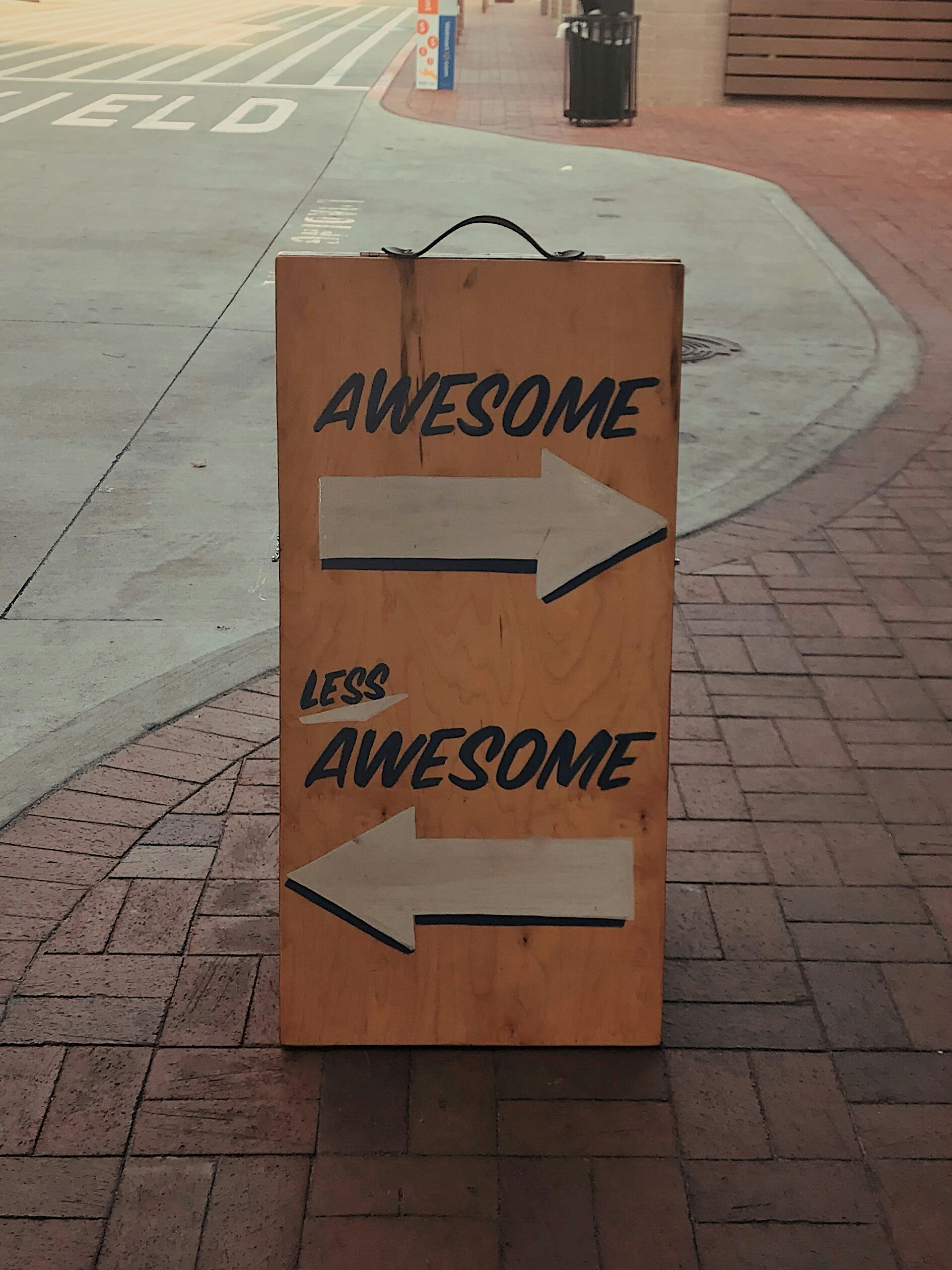 On a brick side walk, a sign has two arrows pointing in opposite directions. One arrow says "Awesome". One arrow says "Less Awesome". 